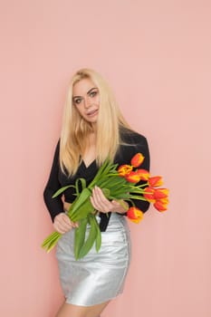 Fashion model woman in fashionable clothes on pink background. Wearing stylish clothing, black blouse, silver skirt. Posing in studio. Holding red tulips in her hands