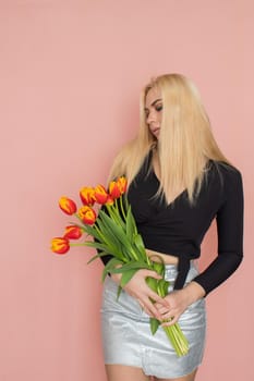 Fashion model woman in fashionable clothes on pink background. Wearing stylish clothing, black blouse, silver skirt. Posing in studio. Holding red tulips in her hands