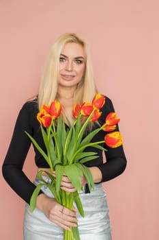 Fashion model woman in fashionable clothes on pink background. Wearing stylish clothing, black blouse, silver skirt. Posing in studio. Holding red tulips in her hands