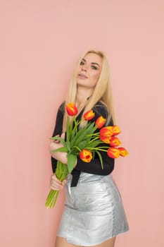 Fashion model woman in fashionable clothes on pink background. Wearing stylish clothing, black blouse, silver skirt. Posing in studio. Holding red tulips in her hands