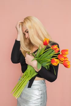 Fashion model woman in fashionable clothes on pink background. Wearing stylish clothing, black blouse, silver skirt. Posing in studio. Holding red tulips in her hands