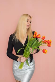 Fashion model woman in fashionable clothes on pink background. Wearing stylish clothing, black blouse, silver skirt. Posing in studio. Holding red tulips in her hands