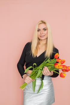 Fashion model woman in fashionable clothes on pink background. Wearing stylish clothing, black blouse, silver skirt. Posing in studio. Holding red tulips in her hands