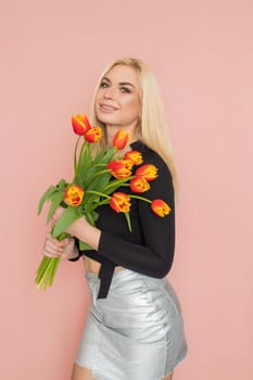 Fashion model woman in fashionable clothes on pink background. Wearing stylish clothing, black blouse, silver skirt. Posing in studio. Holding red tulips in her hands