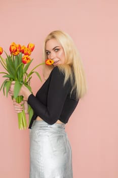 Fashion model woman in fashionable clothes on pink background. Wearing stylish clothing, black blouse, silver skirt. Posing in studio. Holding red tulips in her hands