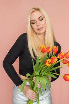 Fashion model woman in fashionable clothes on pink background. Wearing stylish clothing, black blouse, silver skirt. Posing in studio. Holding red tulips in her hands