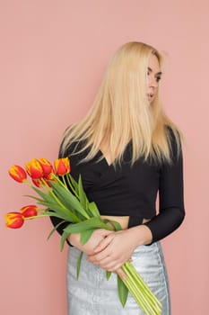 Fashion model woman in fashionable clothes on pink background. Wearing stylish clothing, black blouse, silver skirt. Posing in studio. Holding red tulips in her hands