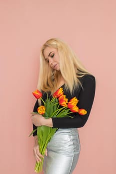 Fashion model woman in fashionable clothes on pink background. Wearing stylish clothing, black blouse, silver skirt. Posing in studio. Holding red tulips in her hands