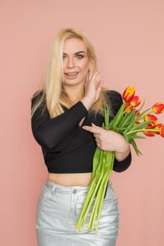 Fashion model woman in fashionable clothes on pink background. Wearing stylish clothing, black blouse, silver skirt. Posing in studio. Holding red tulips in her hands