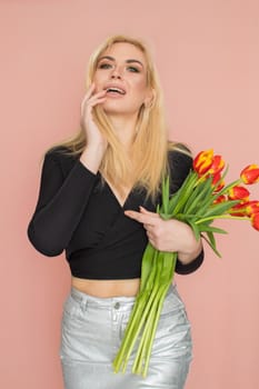 Fashion model woman in fashionable clothes on pink background. Wearing stylish clothing, black blouse, silver skirt. Posing in studio. Holding red tulips in her hands