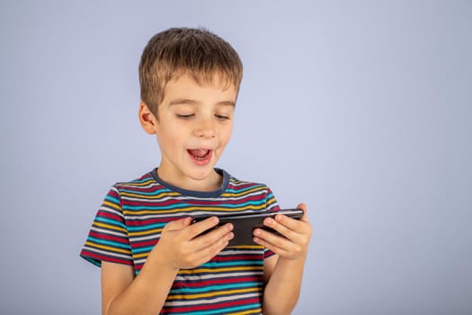 A little boy in a striped T-shirt smiles and looks at the smartphone screen. On a plain blue background with a place to copy.
