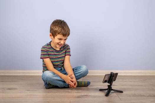 A little boy in a striped T-shirt smiles and looks at the smartphone screen in a stand on the floor. On a plain blue background with a place to copy.