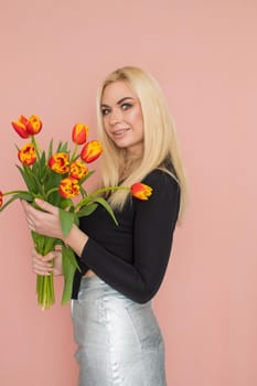 Fashion model woman in fashionable clothes on pink background. Wearing stylish clothing, black blouse, silver skirt. Posing in studio. Holding red tulips in her hands