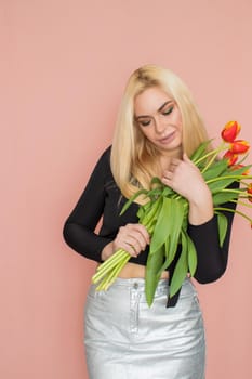 Fashion model woman in fashionable clothes on pink background. Wearing stylish clothing, black blouse, silver skirt. Posing in studio. Holding red tulips in her hands