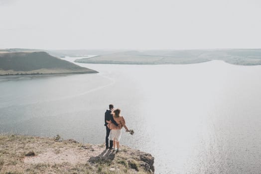The groom and bride in wedding dress stand on a cliff in front of a large reservoir in distance are islands. Warm sunny summer weather. Spring green grass.A girl in a pink jacket holds a bouquet.