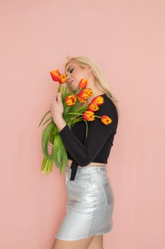 Fashion model woman in fashionable clothes on pink background. Wearing stylish clothing, black blouse, silver skirt. Posing in studio. Holding red tulips in her hands