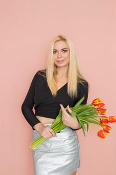 Fashion model woman in fashionable clothes on pink background. Wearing stylish clothing, black blouse, silver skirt. Posing in studio. Holding red tulips in her hands