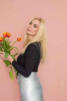 Fashion model woman in fashionable clothes on pink background. Wearing stylish clothing, black blouse, silver skirt. Posing in studio. Holding red tulips in her hands