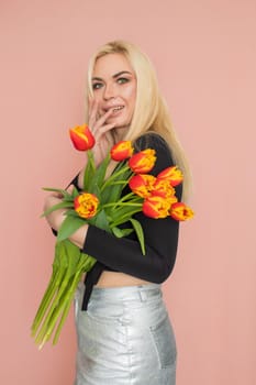 Fashion model woman in fashionable clothes on pink background. Wearing stylish clothing, black blouse, silver skirt. Posing in studio. Holding red tulips in her hands