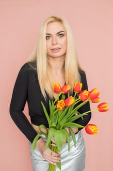 Fashion model woman in fashionable clothes on pink background. Wearing stylish clothing, black blouse, silver skirt. Posing in studio. Holding red tulips in her hands