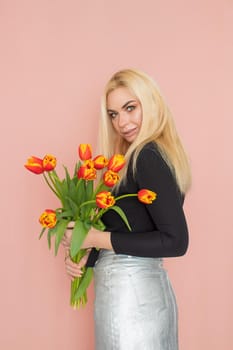 Fashion model woman in fashionable clothes on pink background. Wearing stylish clothing, black blouse, silver skirt. Posing in studio. Holding red tulips in her hands