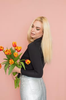 Fashion model woman in fashionable clothes on pink background. Wearing stylish clothing, black blouse, silver skirt. Posing in studio. Holding red tulips in her hands