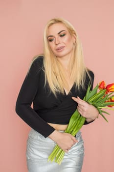 Fashion model woman in fashionable clothes on pink background. Wearing stylish clothing, black blouse, silver skirt. Posing in studio. Holding red tulips in her hands