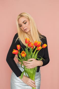 Fashion model woman in fashionable clothes on pink background. Wearing stylish clothing, black blouse, silver skirt. Posing in studio. Holding red tulips in her hands