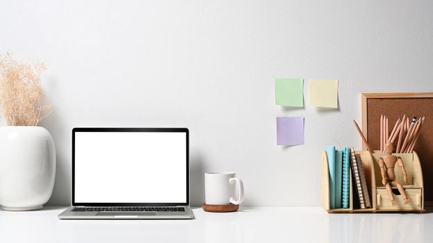 Mockup laptop computer with blank screen, flower pot, coffee cup and stationery on white table. Home office desk.