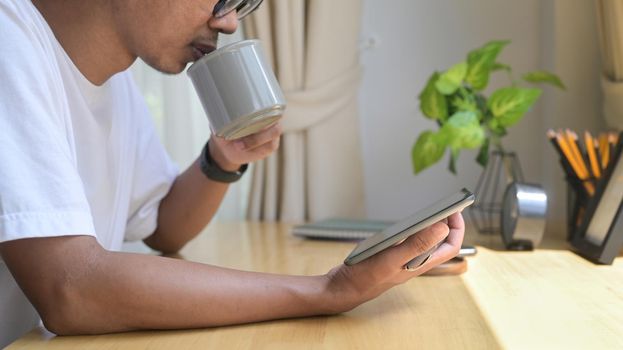 Man drinking hot coffee and reading email, checking news on digital tablet in the morning at home.