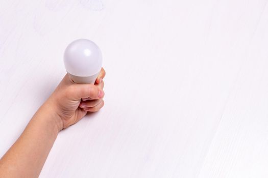 A child's hand holds an LED light bulb on the background of a light wooden table
