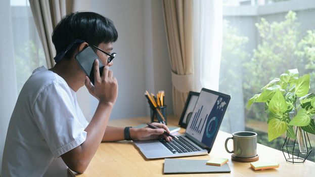 Young man having cell phone conversation and working with computer laptop a home.