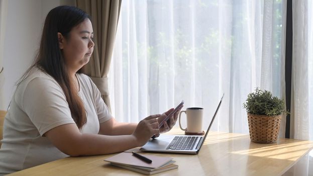 Side view chubby asian woman sitting in front of laptop computer and using mobile phone at home.