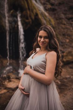 pregnant woman with a big belly in a white long dress in nature on the background of a waterfall