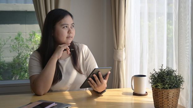 Thoughtful young female freelancer sitting in home office with digital tablet and looking through window.
