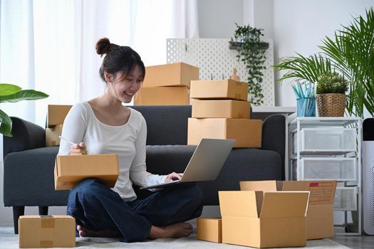 Young business woman answering customers through laptops and preparing parcel boxes of product for delivery. Online selling, e-commerce concept.
