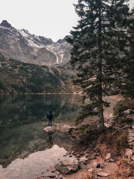 a young man a guy in a hood stands on the Big Stone in the middle of a clear azure green lake surrounded by high mountains and trees. Snow-covered tops of mountains and dry conifers in the fog.