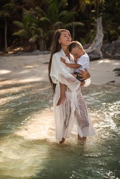 A young tall, long-haired brunette, a fair-haired girl, a mother, is walking along the azure sea and carries a small, fair-haired, happy son in her arms.white cotton clothes. boho dress.