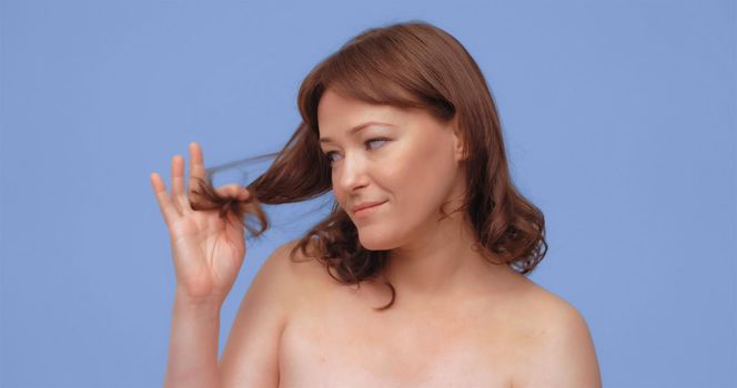 Red-haired woman holds and looks at split ends of her hair. Adult lady with naked shoilders, cut out on blue background. Hair care concept. Close up portrait.