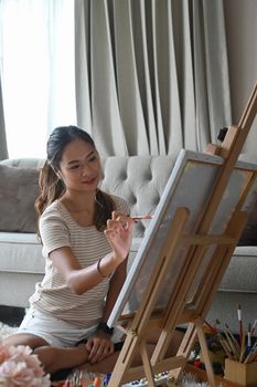 Pretty young woman painting a picture on canvas in bright living room.