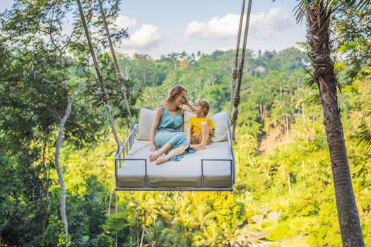 Mother and son swinging in the jungle rainforest of Bali island, Indonesia. Swing in the tropics. Swings - trend of Bali. Traveling with kids concept. What to do with children. Child friendly place.