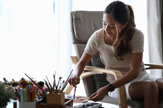 Beautiful woman sitting on armchair in living room and mixing color oil paint on palette.