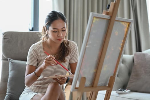Attractive Asian woman painting on canvas at home. Hobby and leisure concept.