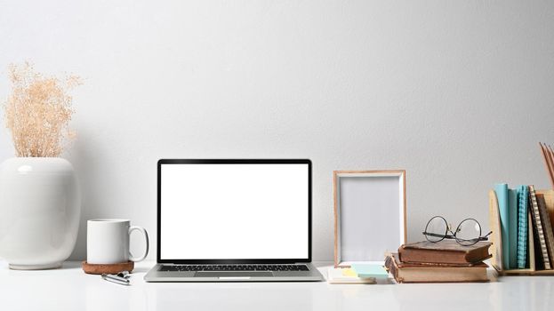 Comfortable workplace with laptop computer, picture frame, flower pot, coffee cup and stationery on white table.