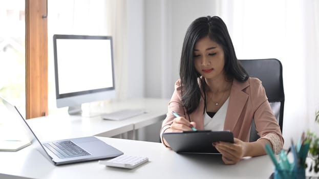 Asian woman sitting at her office desk and writing information on clipboard.