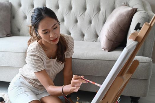 Pretty young woman in casual clothes sitting in living room and painting on easel.