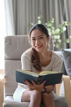 Cheerful millennial woman sitting on armchair in living room and reading book.