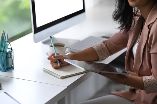 Young businesswoman holding digital tablet and writing information on notebook.