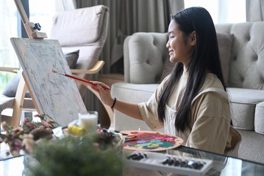 Smiling asian girl painting picture on easel in living room.