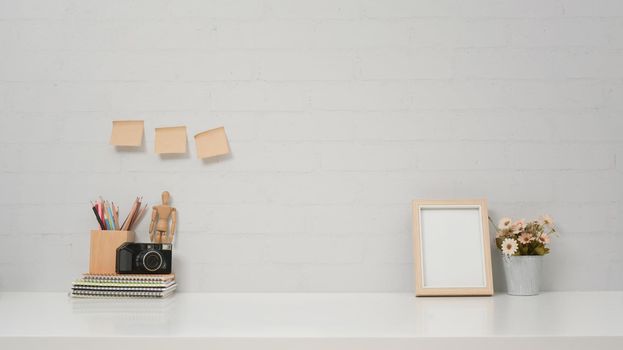 Contemporary workspace with camera, picture frame and flower pot on white table against brick wall.